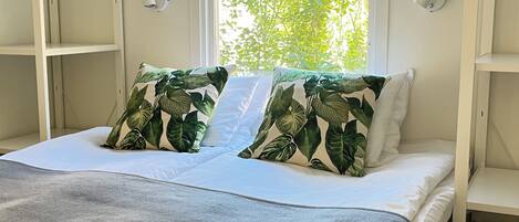A bed with white bedding and two pillows with a tropical leaf pattern, a window with a view of green foliage, and a white wall with a security camera.