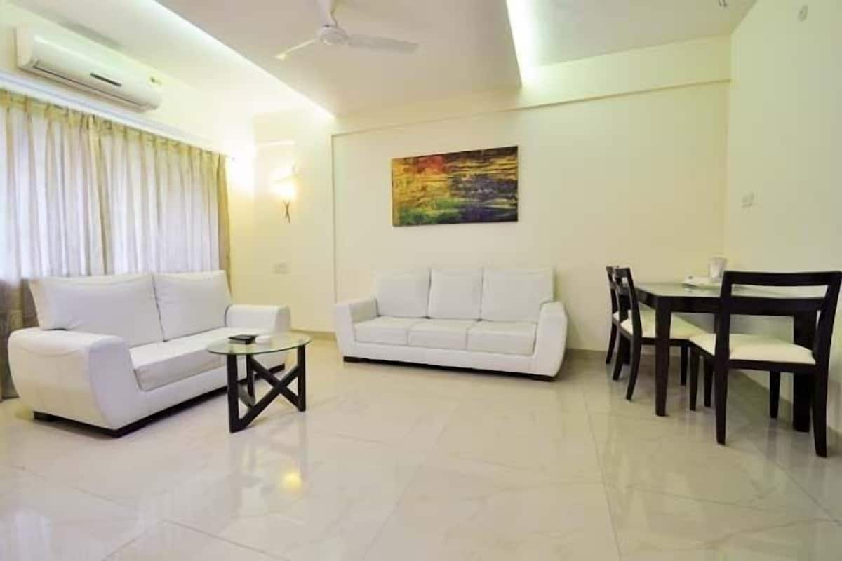 A modern living room with a white sofa, a glass coffee table, a dining table with chairs, and a painting on the wall.