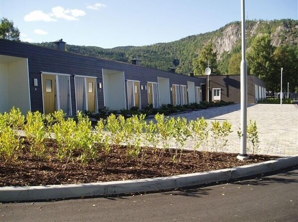 Una hilera de edificios modernos de una sola planta, con diseños de puertas uniformes, un sendero pavimentado y una zona ajardinada con plantas jóvenes.