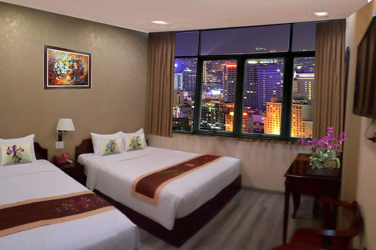 A hotel room with two beds, a large window with a city view, a painting on the wall, and a desk with a chair.