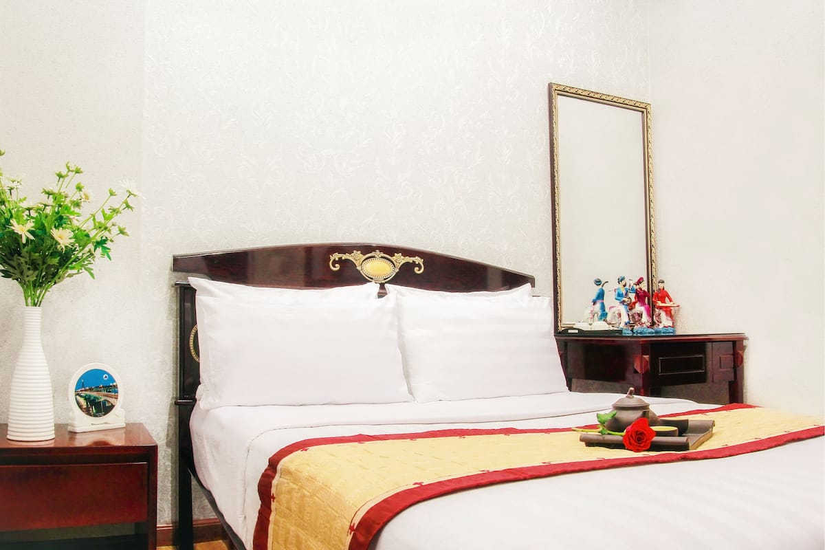 A neatly made bed with white linens and a red and gold bedspread, a wooden nightstand with a vase of flowers, a round mirror, and a small statue.