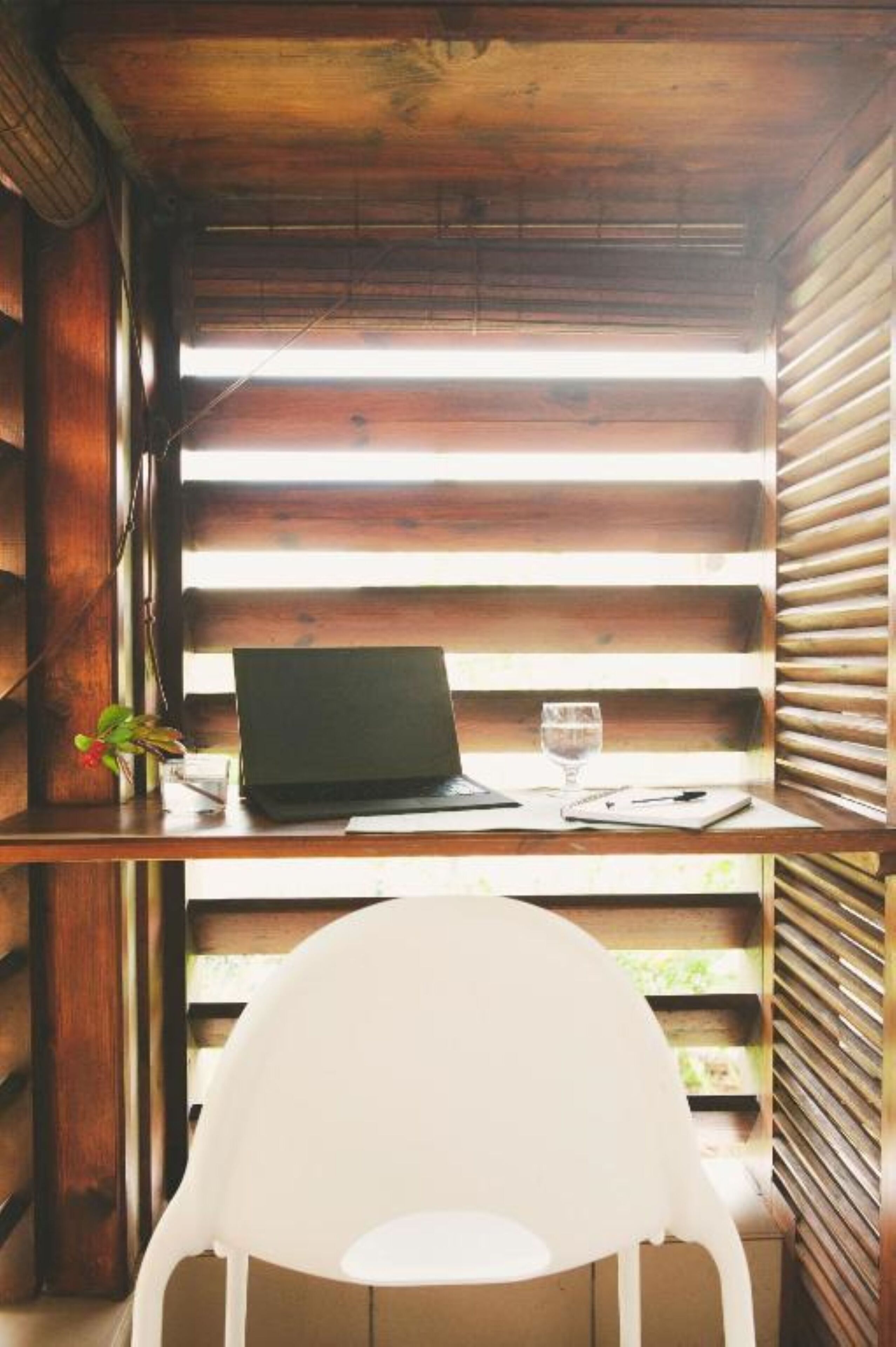 Un bureau en bois avec un ordinateur portable, un verre d’eau et une chaise blanche, dans une pièce dont les murs sont recouverts de lambris en bois.