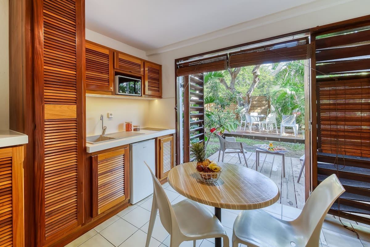 Una cocina compacta con armarios de madera y un refrigerador pequeño. Hay un comedor con una mesa redonda y dos sillas, y una vista a un jardín a través de una puerta corrediza.
