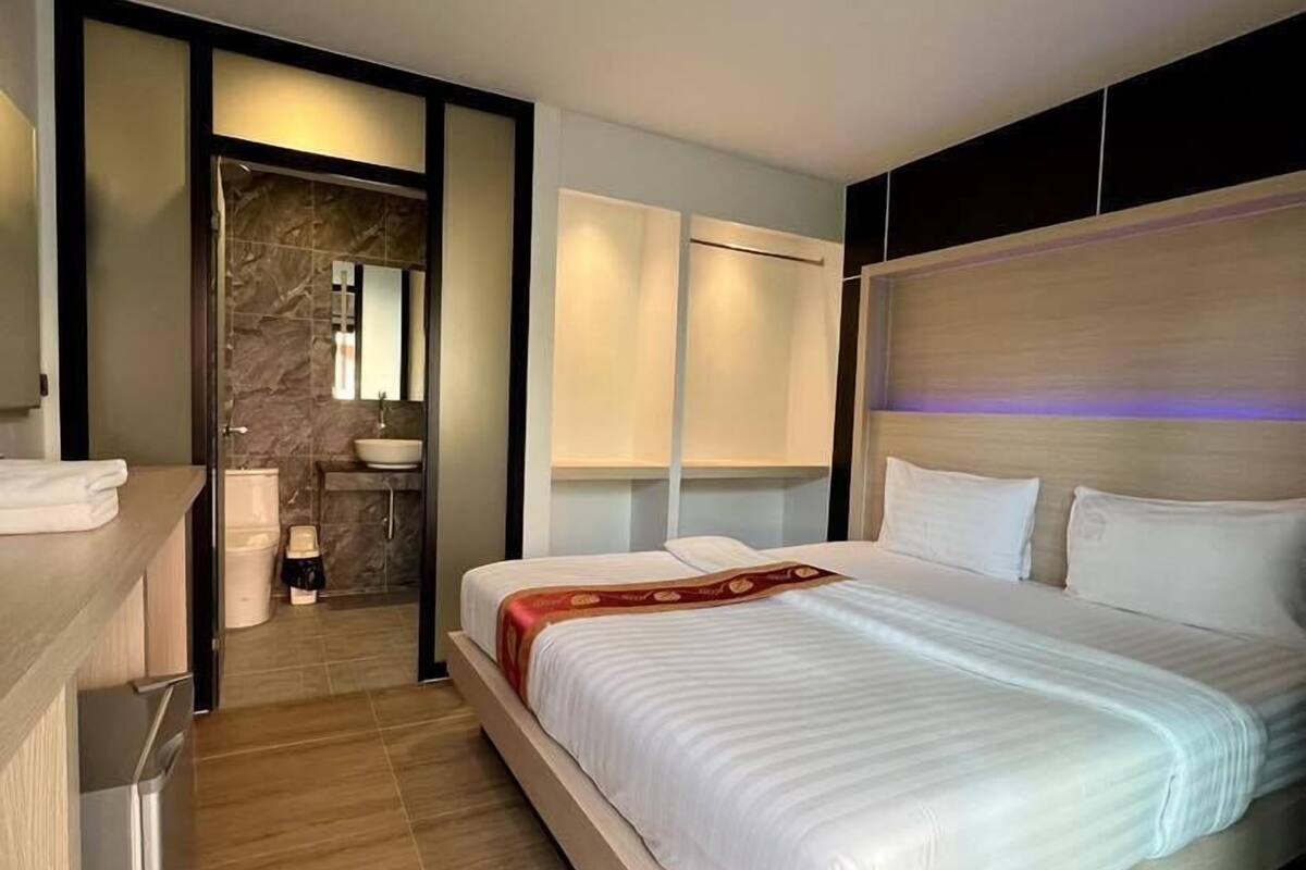 Una habitación de hotel moderna con una cama grande, una mesita de noche, un banco empotrado y un baño con pared y lavamanos de mármol.