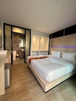 Una habitación de hotel moderna con una cama grande, una mesita de noche, un banco empotrado y un baño con pared y lavamanos de mármol.