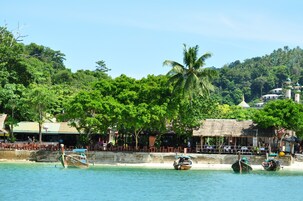 A beachside location with thatched-roof buildings, palm trees, and boats on the water.