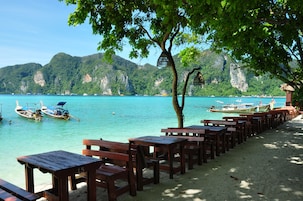 A beach with wooden benches and tables, boats on the water, and a mountainous backdrop.