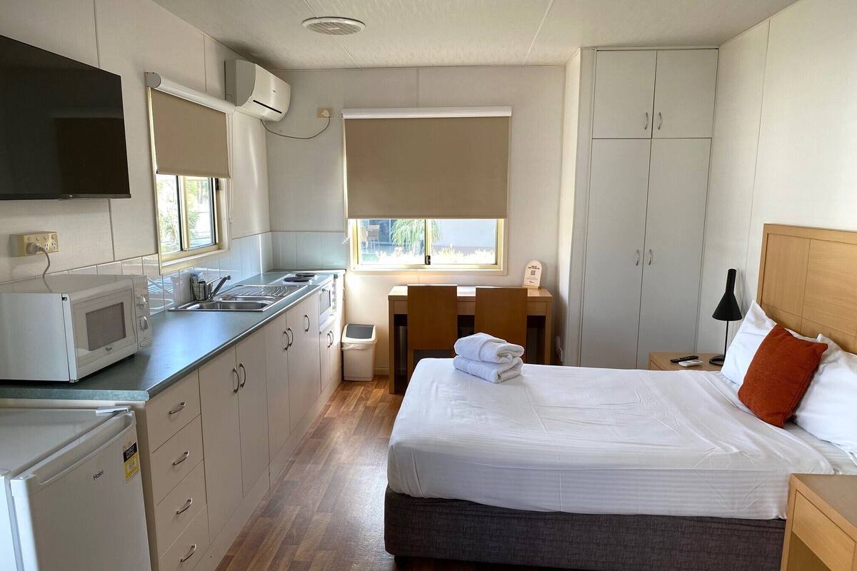 Habitación de hotel pequeña con kitchenette, una cama con almohadas blancas y naranja, una mesita de noche de madera y una ventana con persianas.