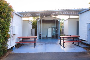 A covered outdoor area with two picnic tables, a metal roof, and a white building in the background.