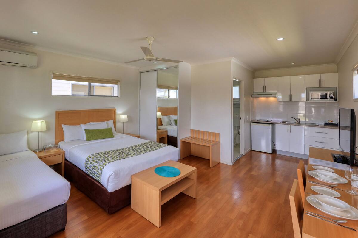 Habitación de hotel con dos camas, un banco de madera, una mesita, kitchenette y un baño visible a través de una puerta abierta.