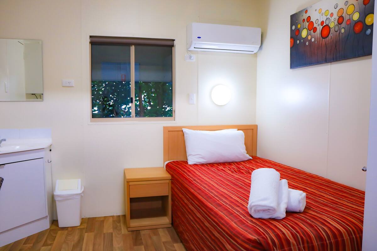 Habitación de hotel con cama, mesita de noche, un cubo de basura pequeño, una ventana con vistas a árboles y un equipo de aire acondicionado.