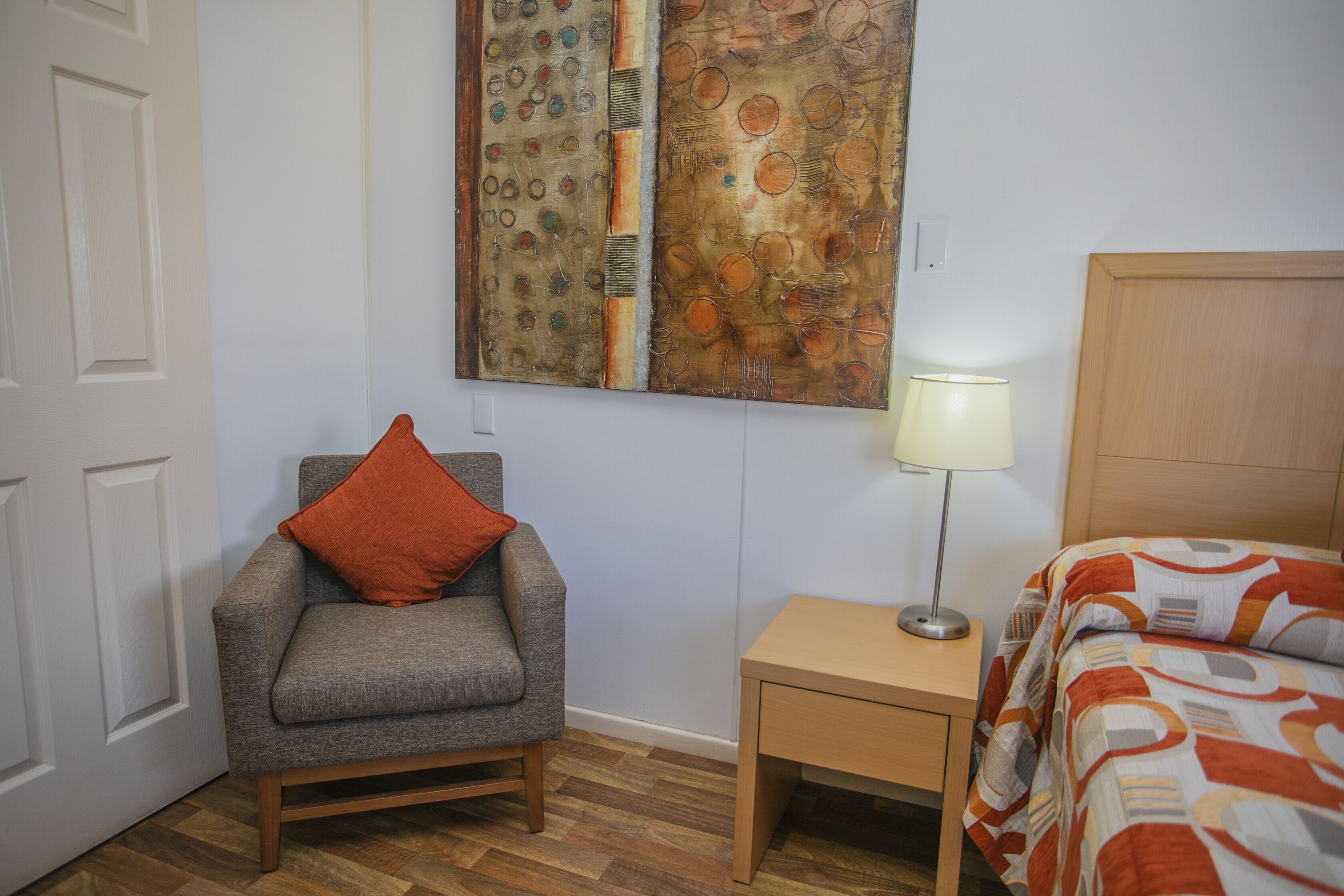 A room with a grey armchair, a wooden nightstand with a lamp, and a bed with patterned bedding.