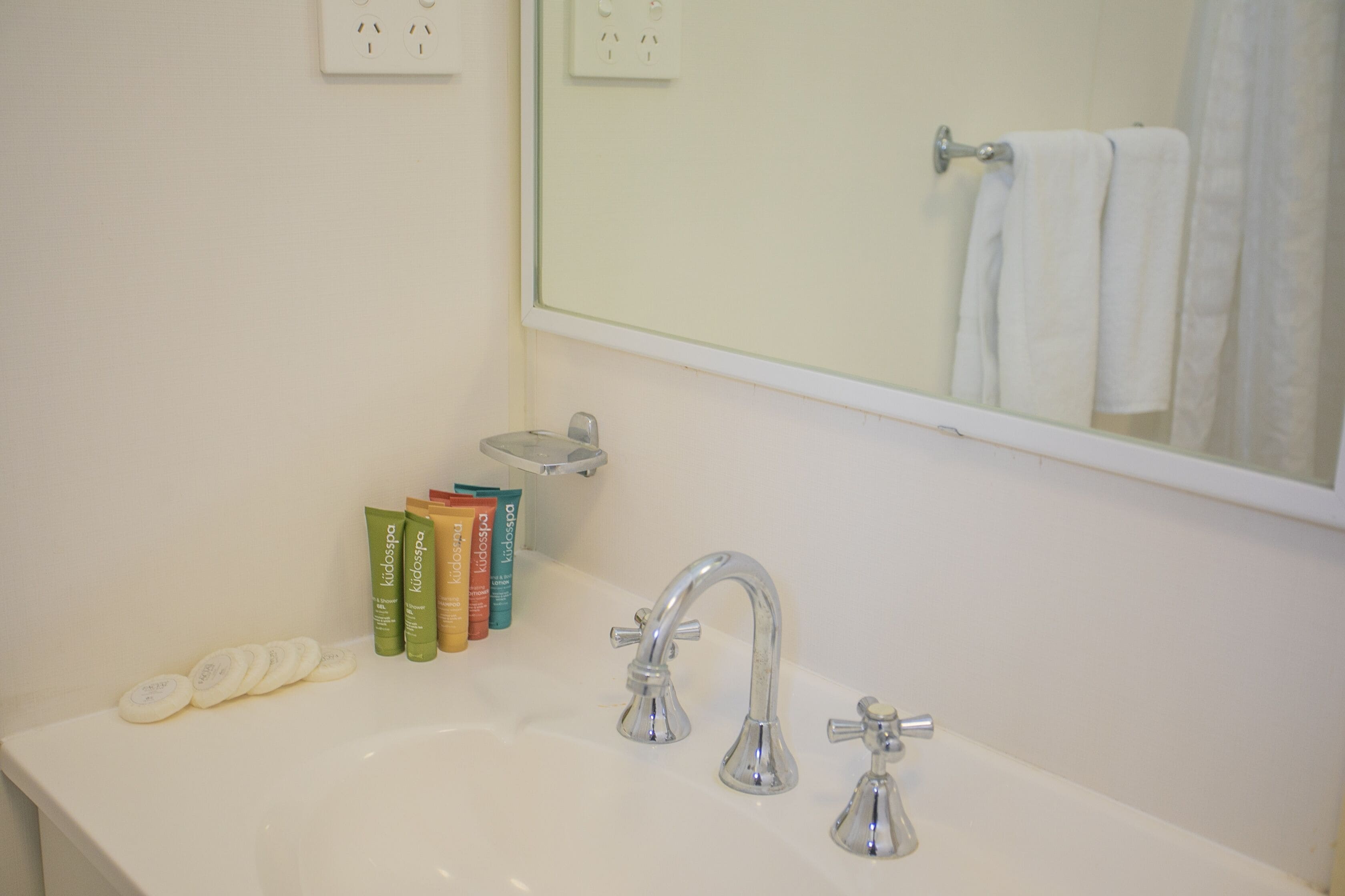 A bathroom with a white sink, two faucets, and a mirror above it. There are toiletries on the sink and a towel hanging on the wall.