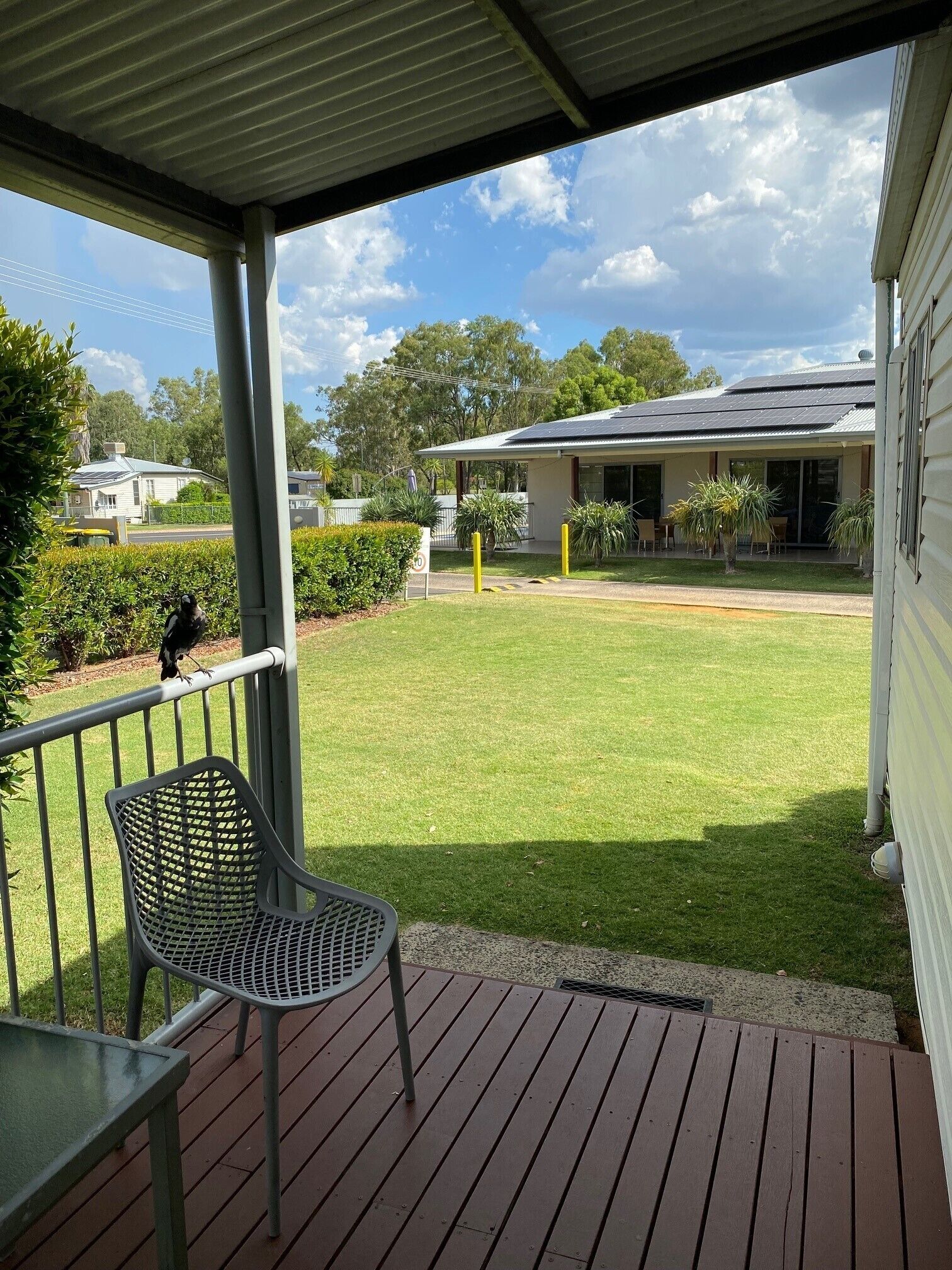 A covered outdoor area with a metal chair, a grassy lawn, and a building with a solar panel roof in the background.