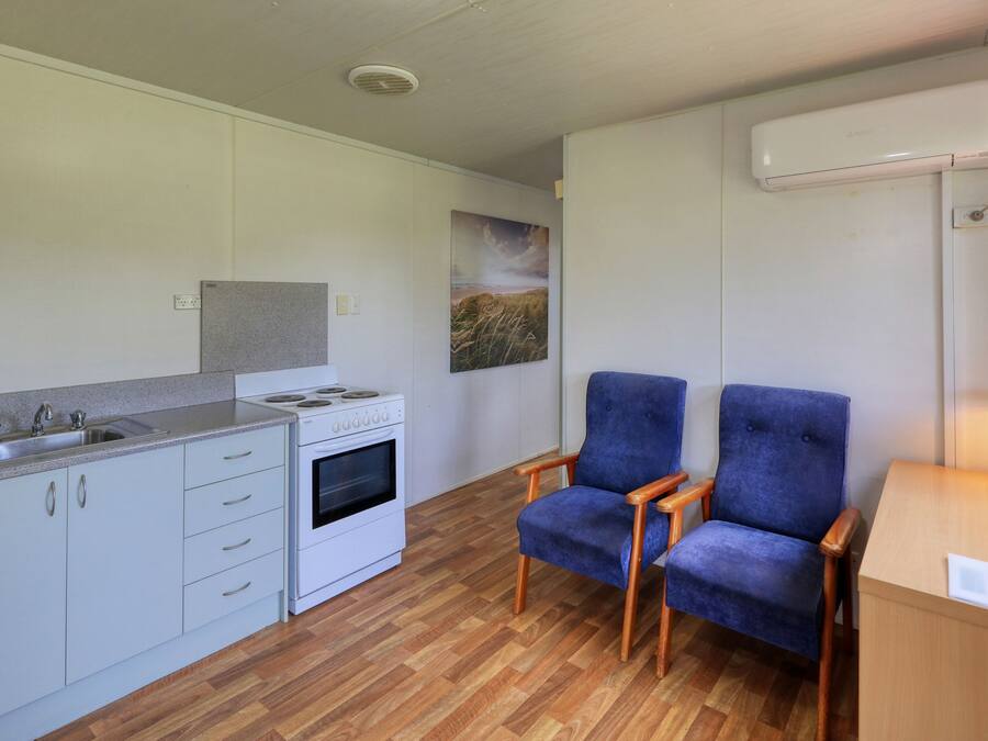 A compact kitchen with a stove, sink, and cabinets. There are two blue chairs and a small table with a lamp.