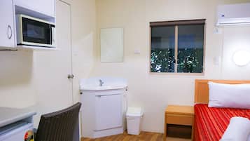 A compact hotel room with a bed, a small bedside table, a microwave, a sink, and a window with a view of trees.