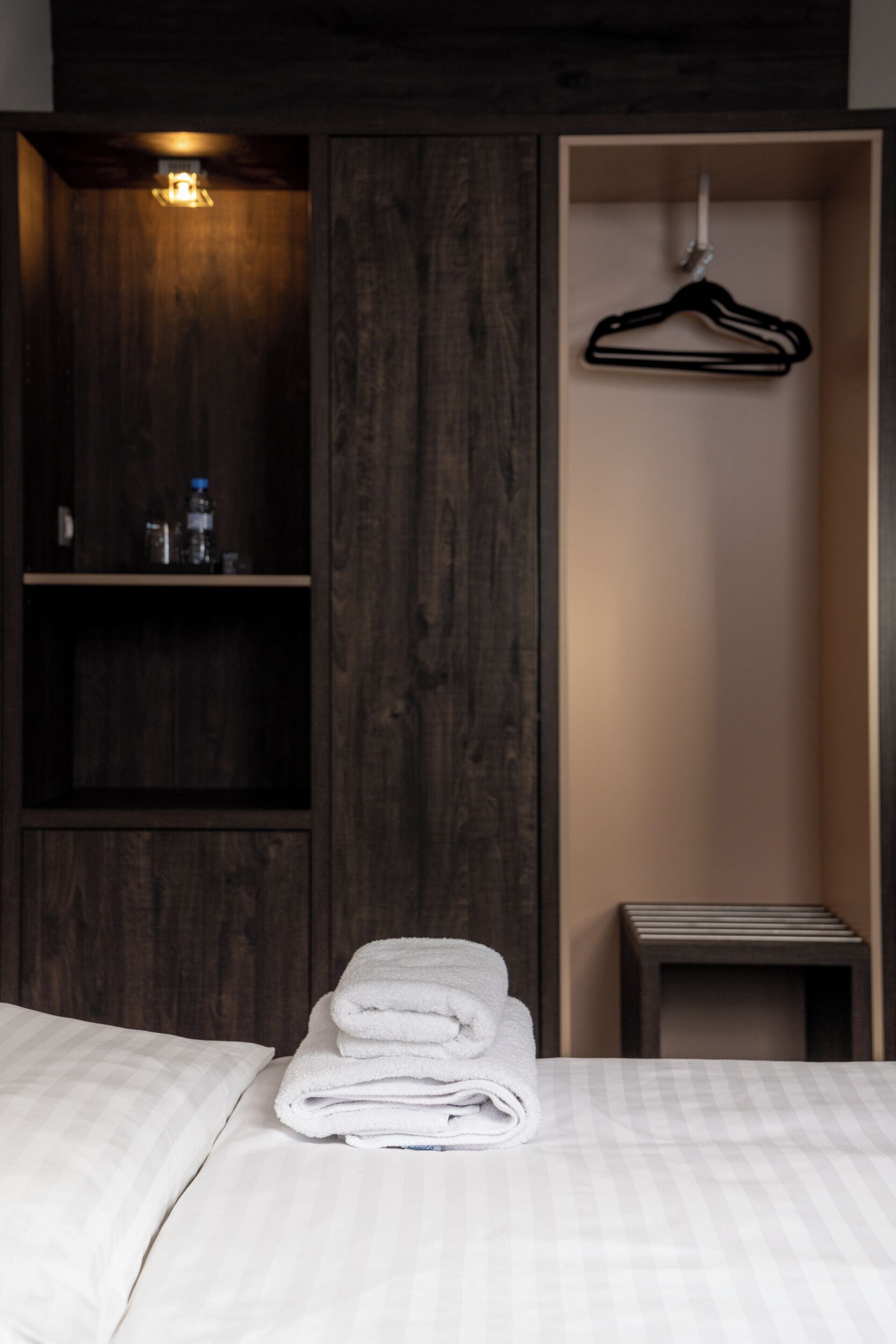 Uma cama bem arrumada com lençóis brancos, um guarda-roupa de madeira com cabideiro e uma pequena mesa de cabeceira.