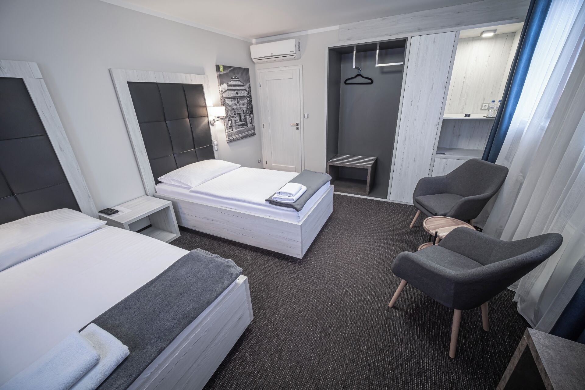 A hotel room with a bed, two chairs, a small table, a wardrobe, and a bathroom with a shower and toilet.