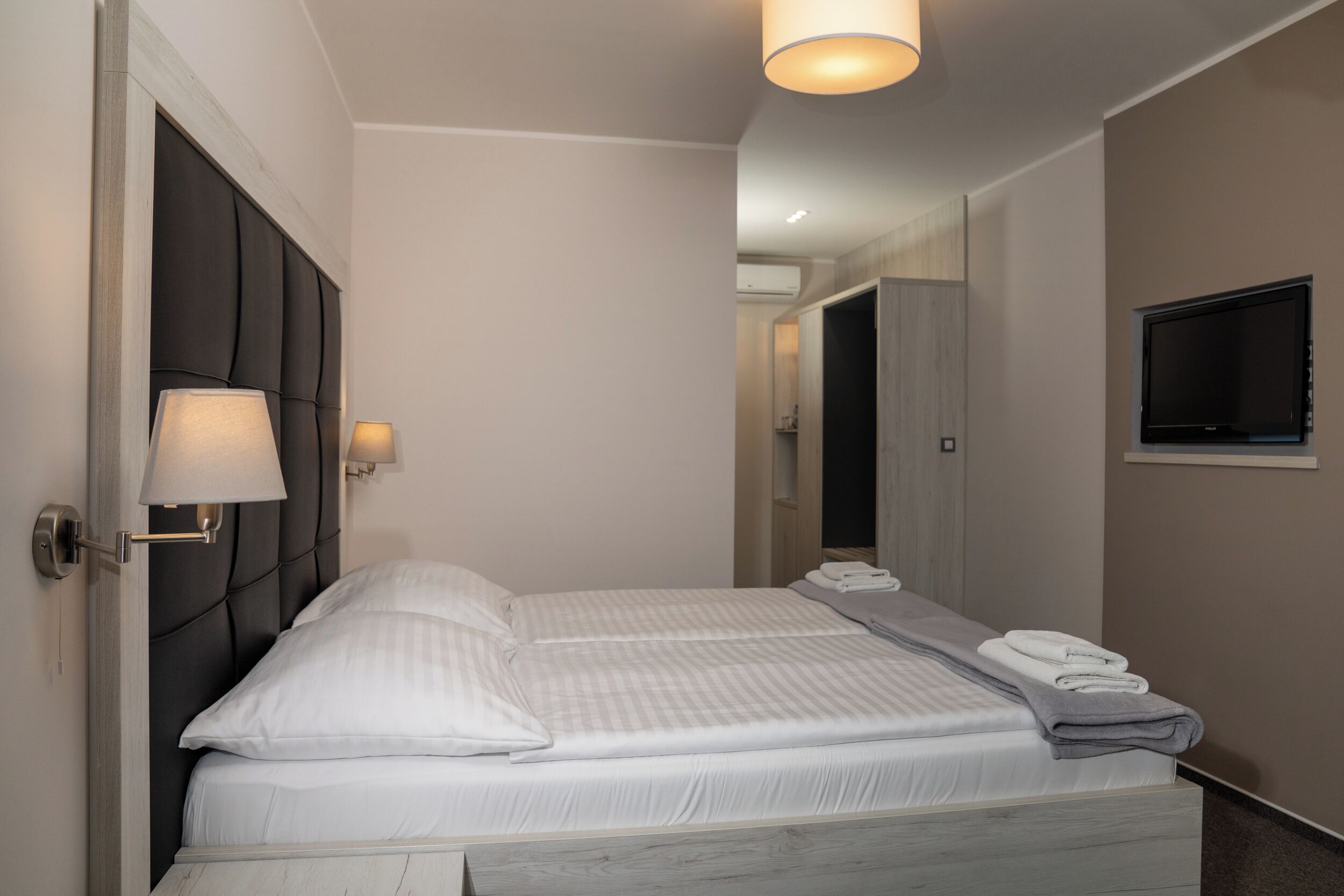 A modern hotel room with a large bed, a bedside lamp, a flat-screen TV, and a neatly made bed with towels.