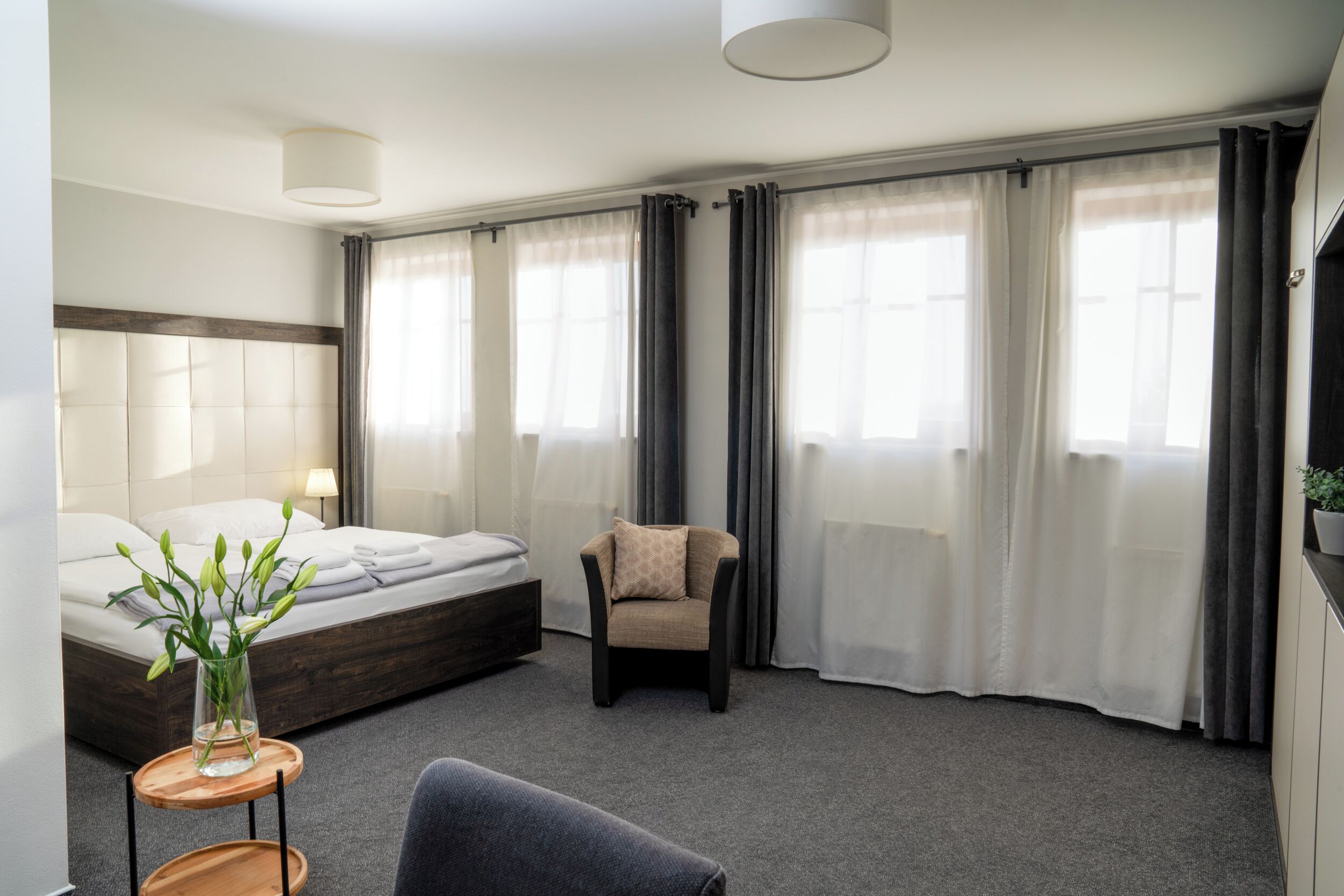 A modern hotel room with a bed, a chair, a small table with a vase, and large windows with curtains.
