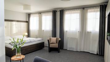 A modern hotel room with a bed, a chair, a small table with a vase, and large windows with curtains.