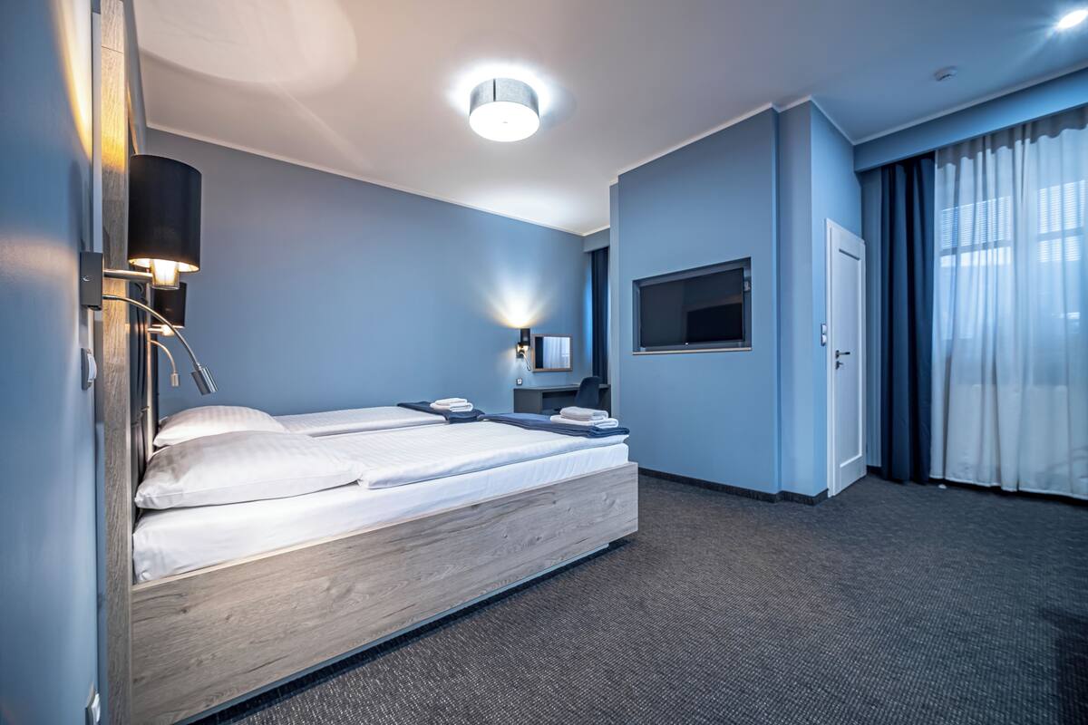 A hotel room with a wooden bed, a flat-screen TV, a desk with a chair, and a large window with curtains.