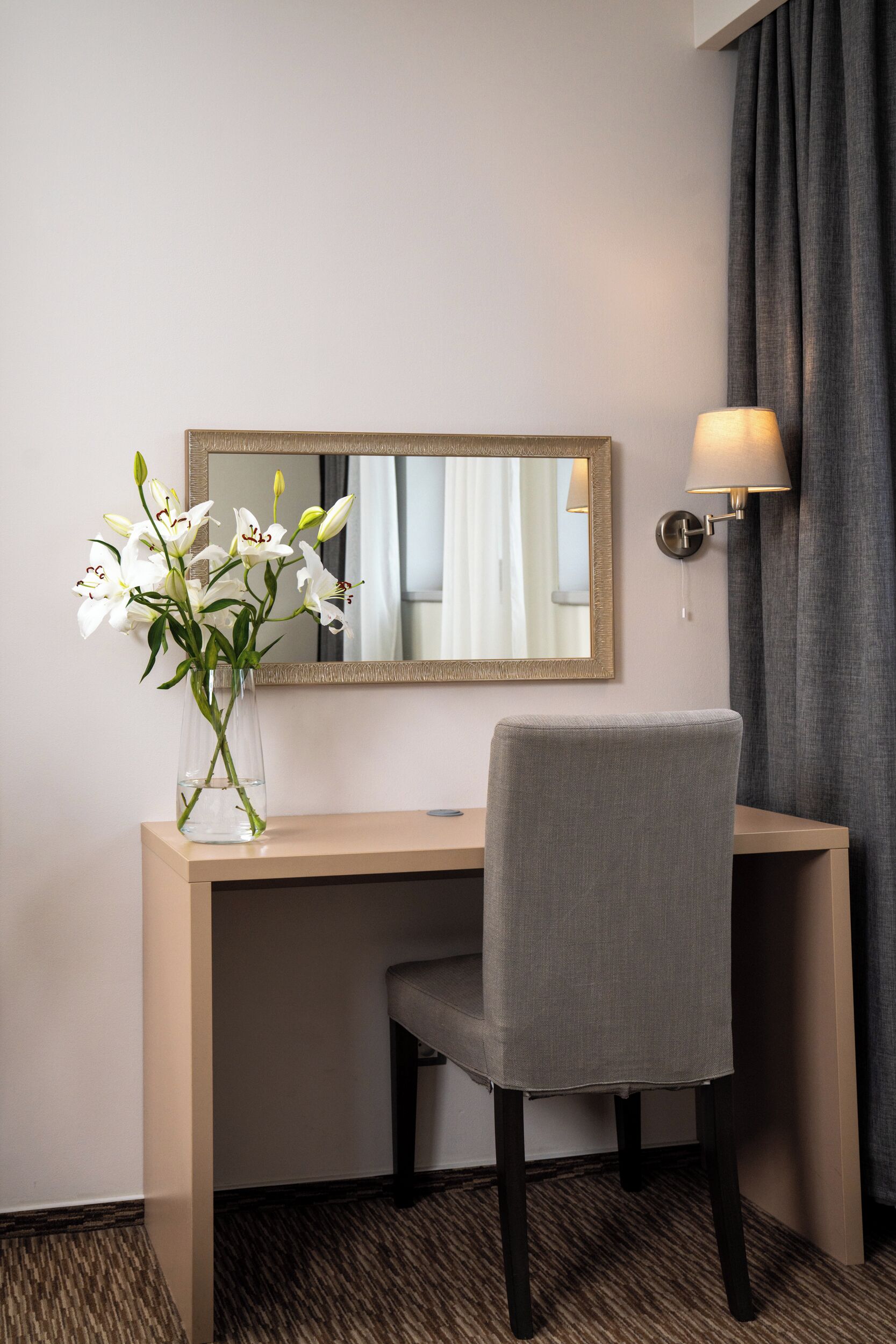 A hotel room with a desk, a chair, a vase with white flowers, a mirror, and a wall-mounted lamp.