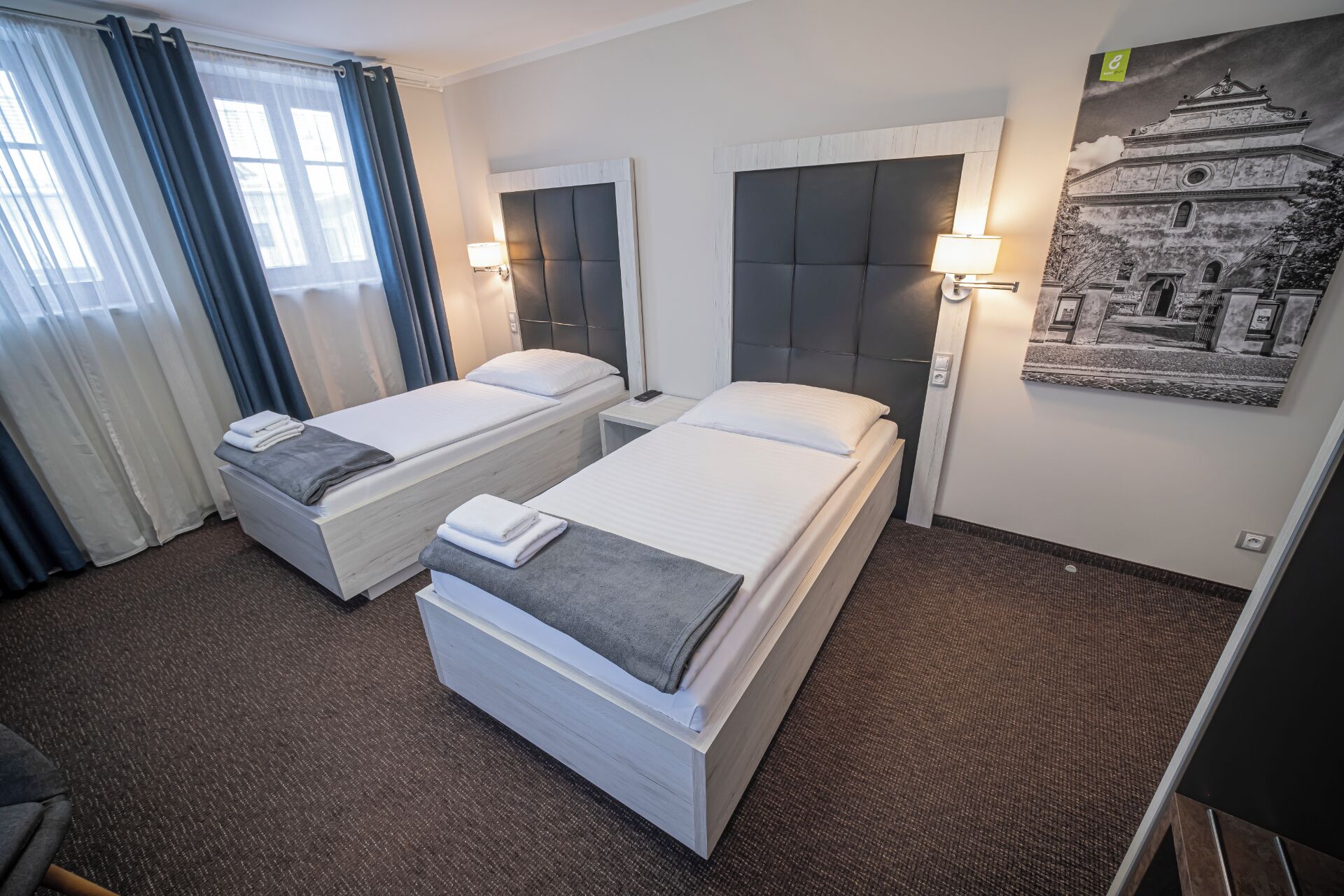 A hotel room with two single beds, a desk, a chair, a window with curtains, and a wall-mounted lamp.
