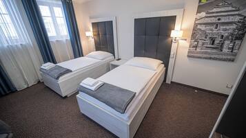 A hotel room with two single beds, a desk, a chair, a window with curtains, and a wall-mounted lamp.