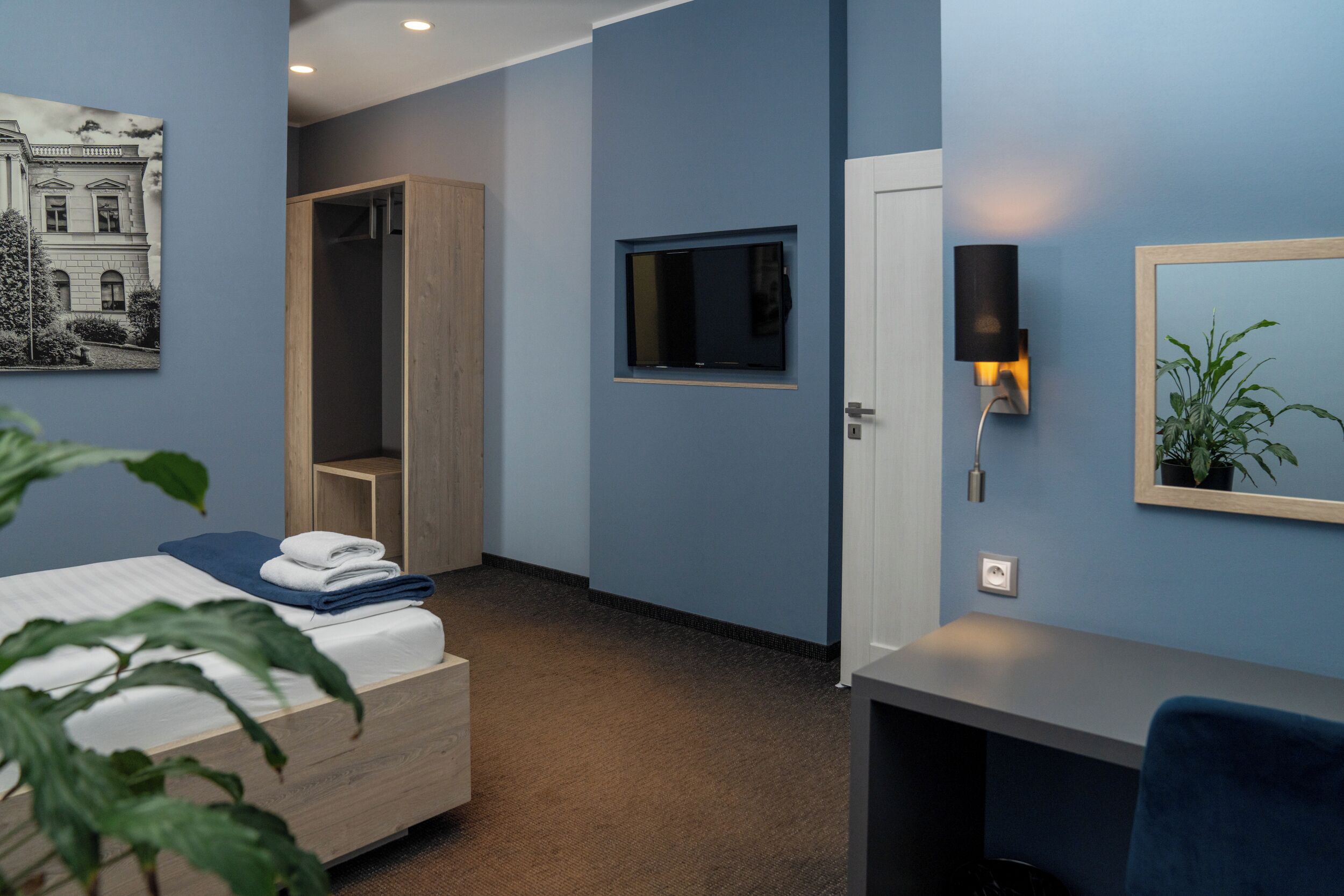 A hotel room with a bed, a wooden wardrobe, a desk, a chair, a TV mounted on the wall, and a wall-mounted lamp.