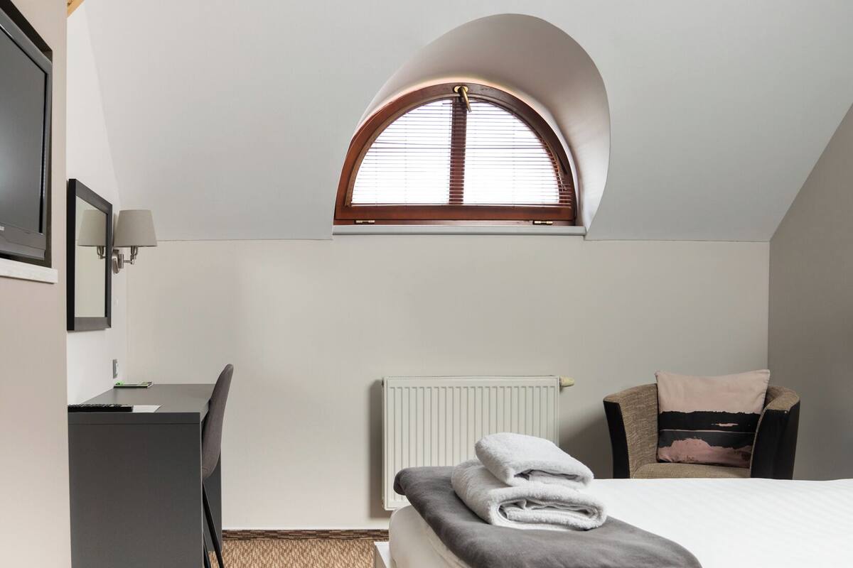 A bedroom with a bed, a desk, a chair, a radiator, and a window with a wooden frame.