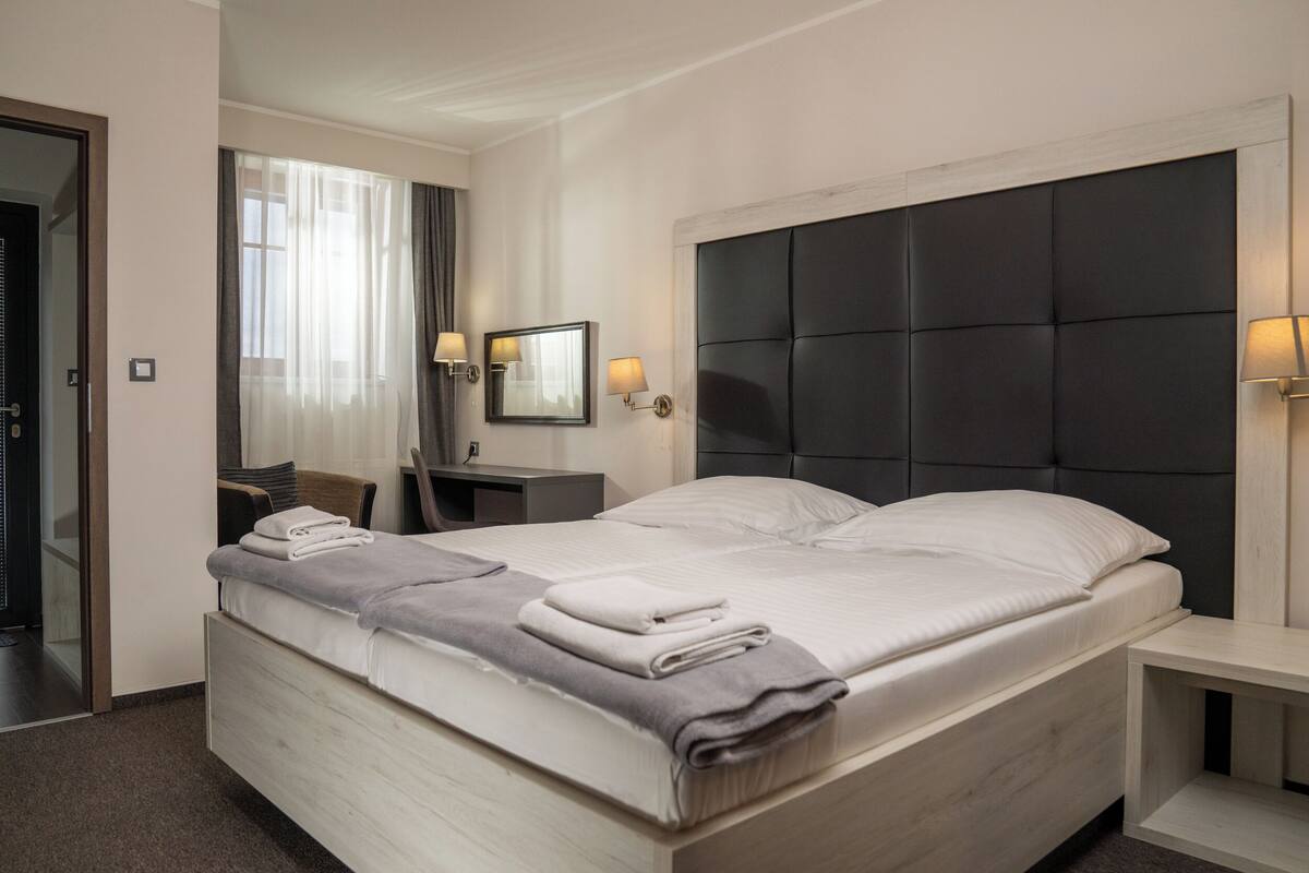 A hotel room with a large bed, a bedside table, a mirror, and a window with curtains.