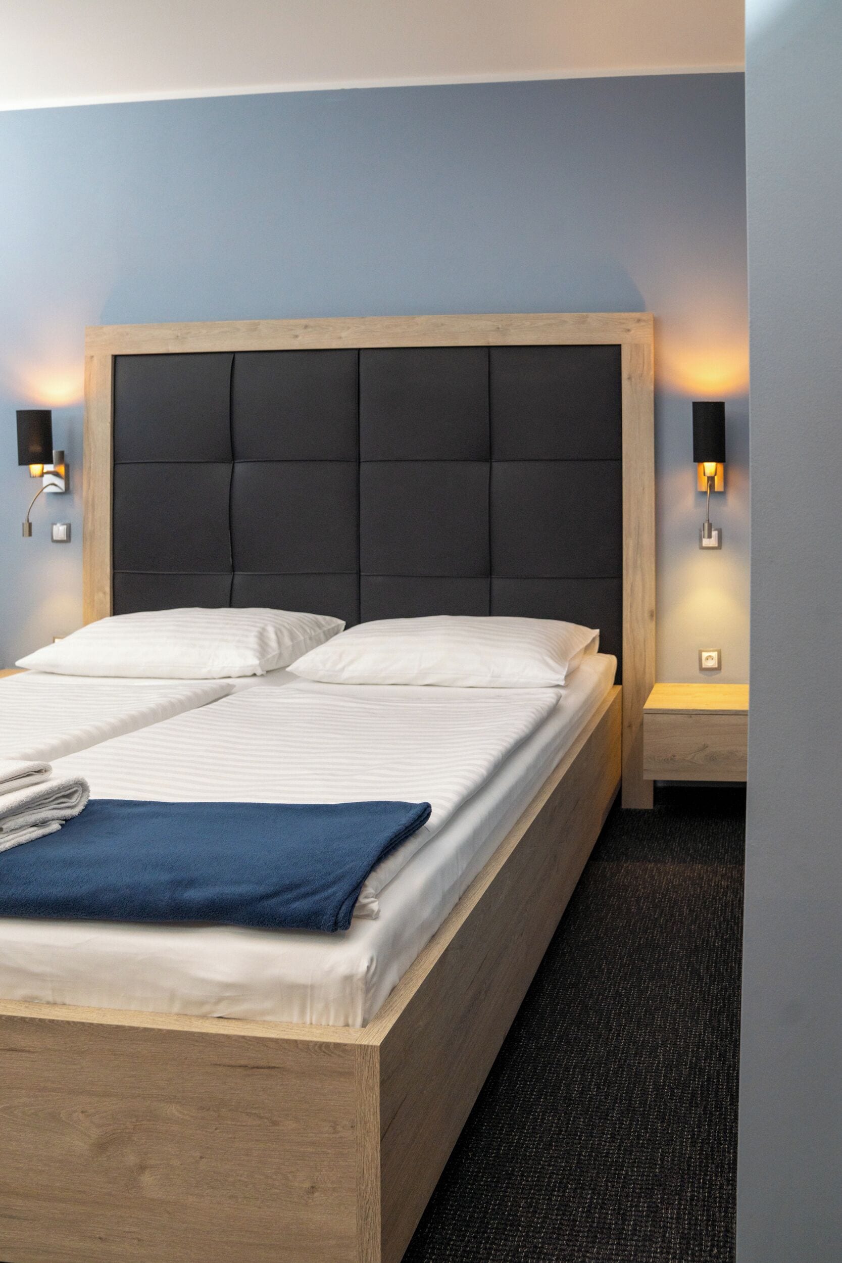Una cama bien hecha con estructura de madera y cabecera oscura, acompañada de dos mesitas de noche con lámparas fijadas a la pared.