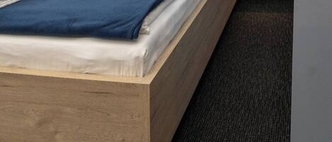 Una cama bien hecha con estructura de madera y cabecera oscura, acompañada de dos mesitas de noche con lámparas fijadas a la pared.