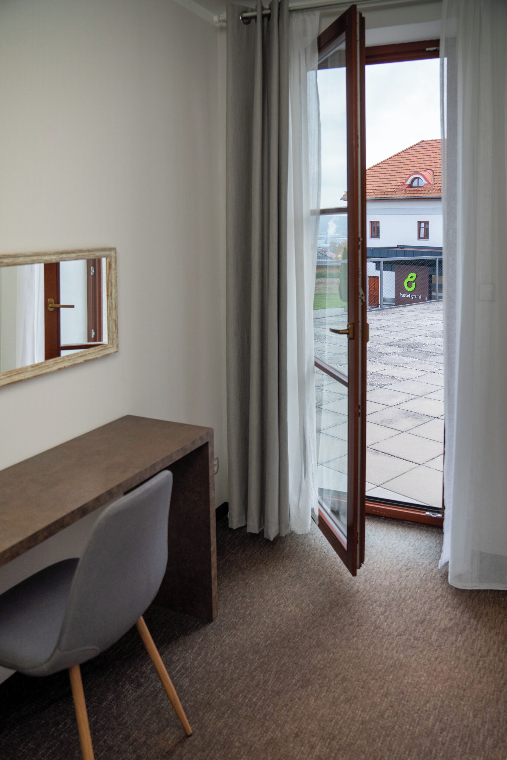 A hotel room with a wooden desk, a chair, a mirror, and a sliding glass door leading to an outdoor area with a building in the background.