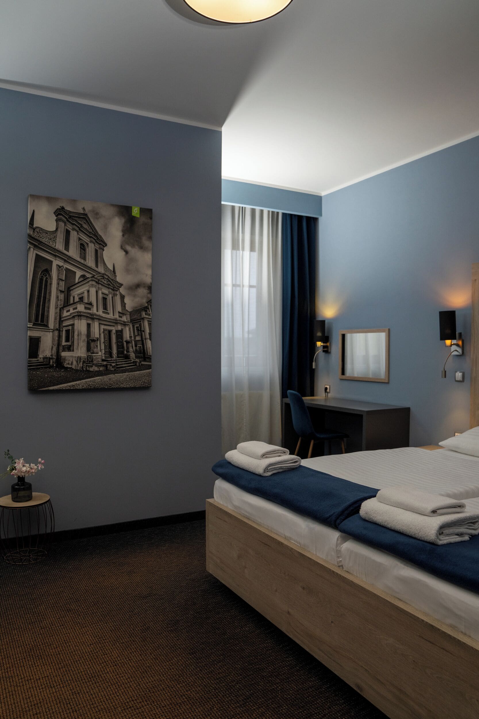 A hotel room with a bed, a desk, a chair, a wall-mounted lamp, a framed picture, and a small table with a vase of flowers.