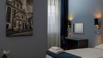 A hotel room with a bed, a desk, a chair, a wall-mounted lamp, a framed picture, and a small table with a vase of flowers.