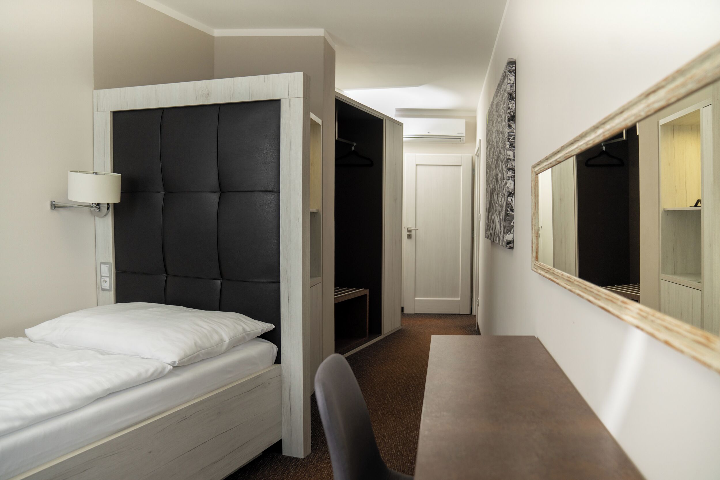 Una habitación de hotel pequeña con cama, escritorio, silla, armario y espejo.