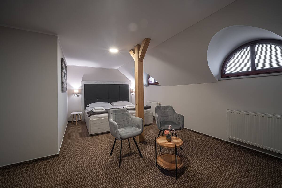 A compact, modern room with a bed, two chairs, a small table, and a large window with blinds.