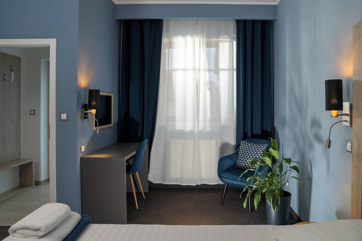 A modern hotel room with a bed, a desk, a chair, a potted plant, and a large window with curtains.