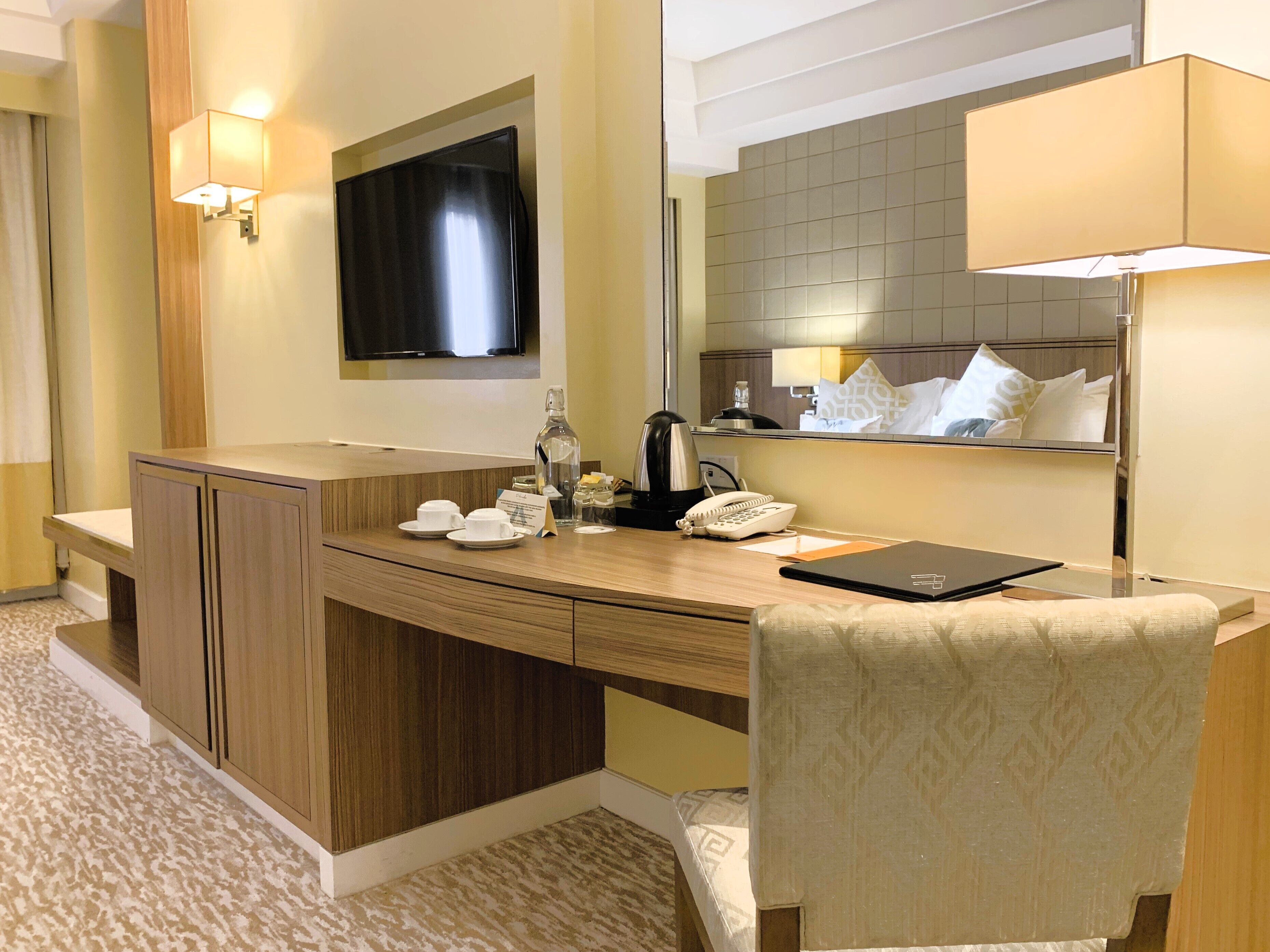 Quarto de hotel com escrivaninha de madeira, uma cadeira, televisão na parede, espelho e cama com colcha estampada.