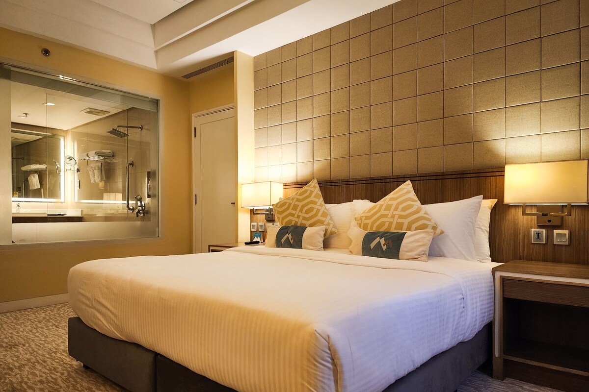 A hotel room with a large bed, a bedside table, a nightstand, a mirror, and a headboard with a light panel.