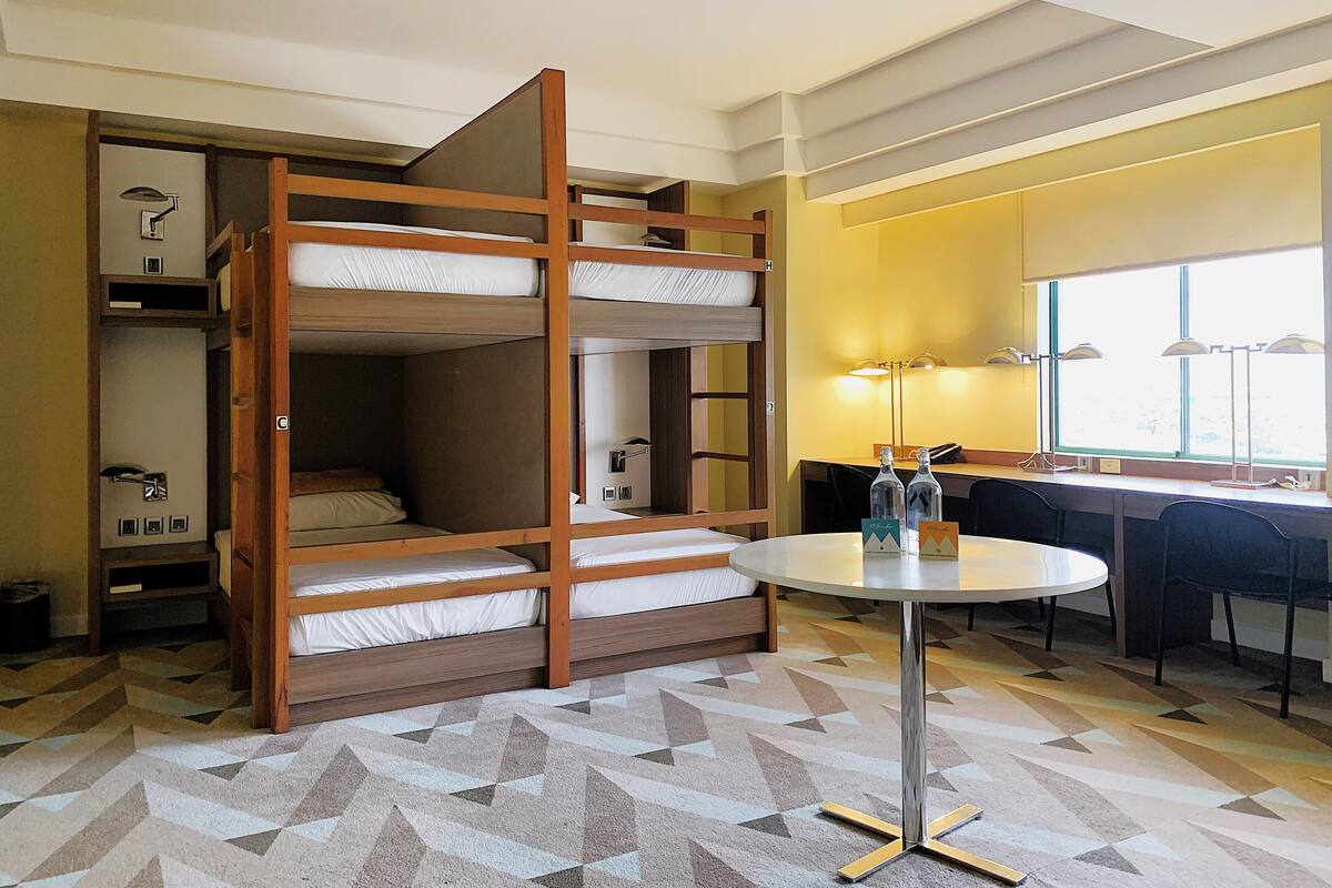Ein Zimmer mit Etagenbetten, einem runden Tisch mit Stühlen, einem Schreibtisch und einem Fenster mit Lampen.