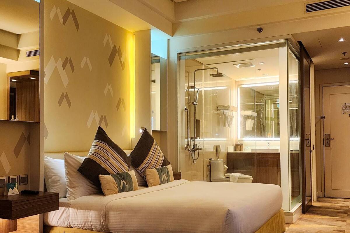 Ein modernes Hotelzimmer mit einem großen Bett, einer gläsernen Dusche und einem sichtbaren Badezimmer mit Spiegel und Handtuchhalter.