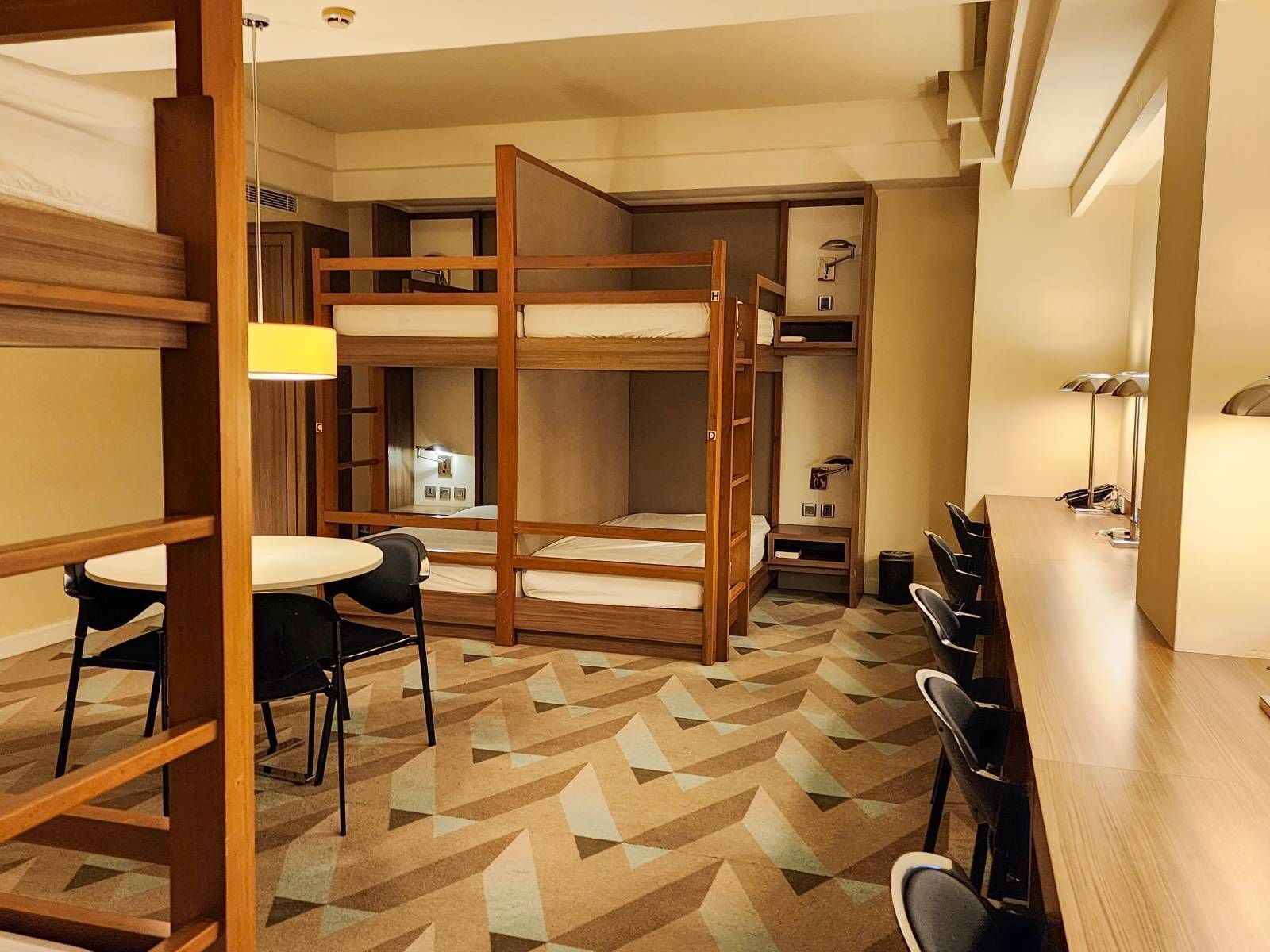 A dormitory room with bunk beds, a desk, chairs, and a patterned carpet.