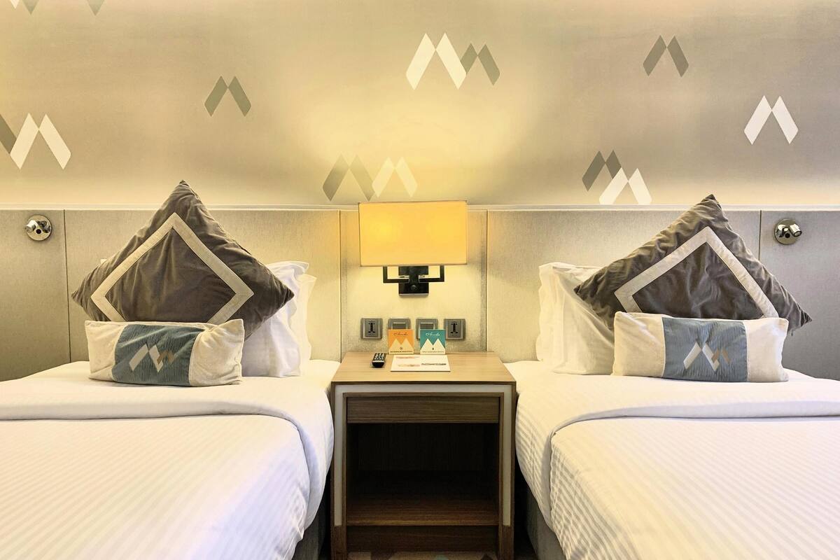 Ein Hotelzimmer mit zwei Betten, jedes mit einem Kissen mit geometrischem Muster. Ein Nachttisch mit Lampe, Fernbedienung und einer Karte. Das Zimmer hat eine neutrale Farbgestaltung und eine strukturierte Wand.