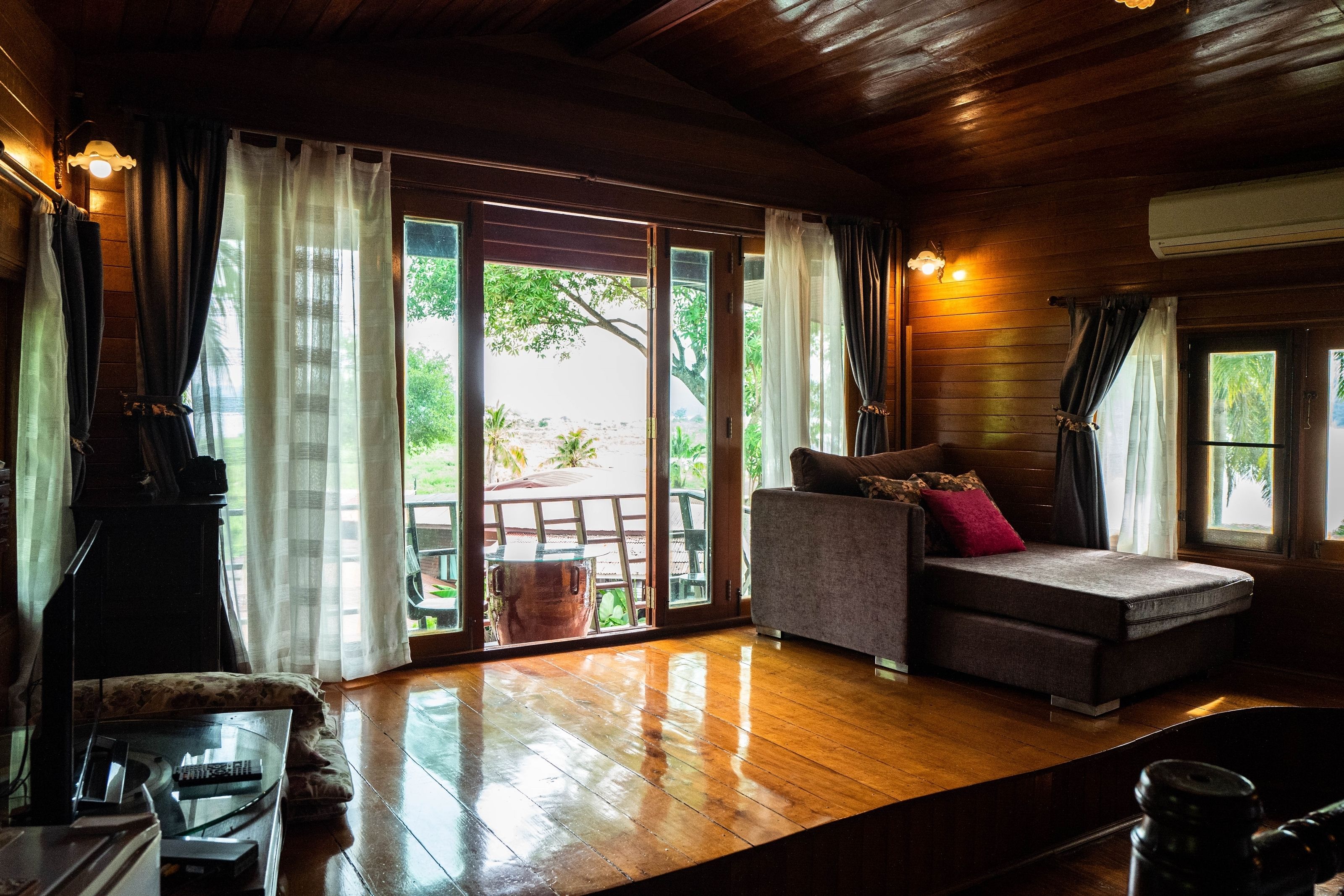 A room with a wooden floor, a sofa with cushions, a television, and a large window with curtains.