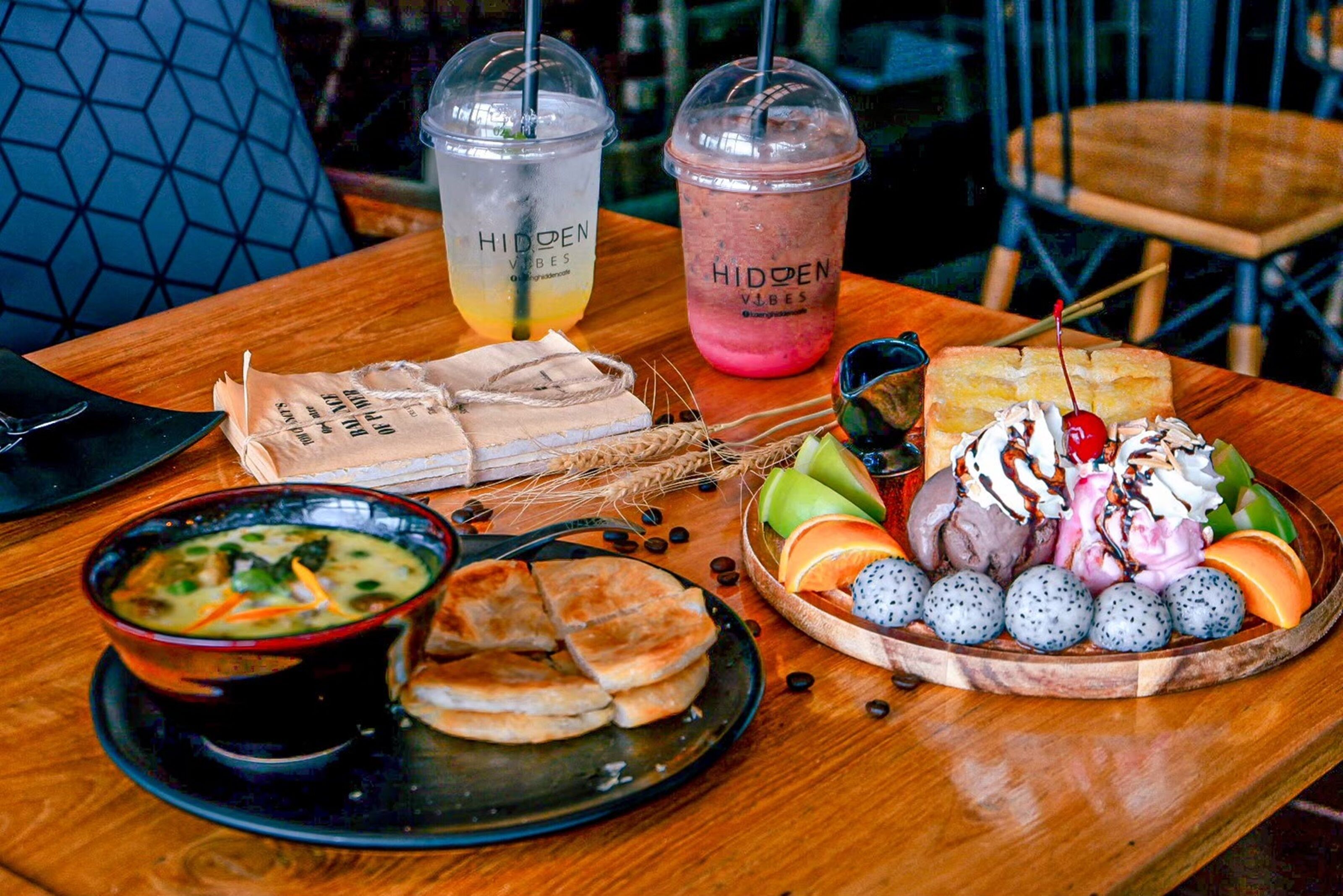 Una mesa con un tazón de sopa, un plato de panqueques, un tazón de fruta y dos bebidas que dicen 'HIDDEN VIBES'.