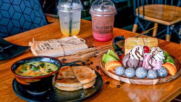 Una mesa con un tazón de sopa, un plato de panqueques, un tazón de fruta y dos bebidas que dicen 'HIDDEN VIBES'.