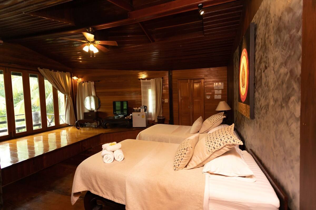 Habitación con dos camas, ventilador de techo, televisor y vista a palmeras.